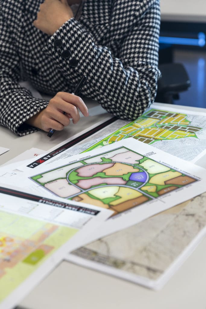 Neck down image of person in jacket leaning over a desk with landscaping plans spread over desk.
